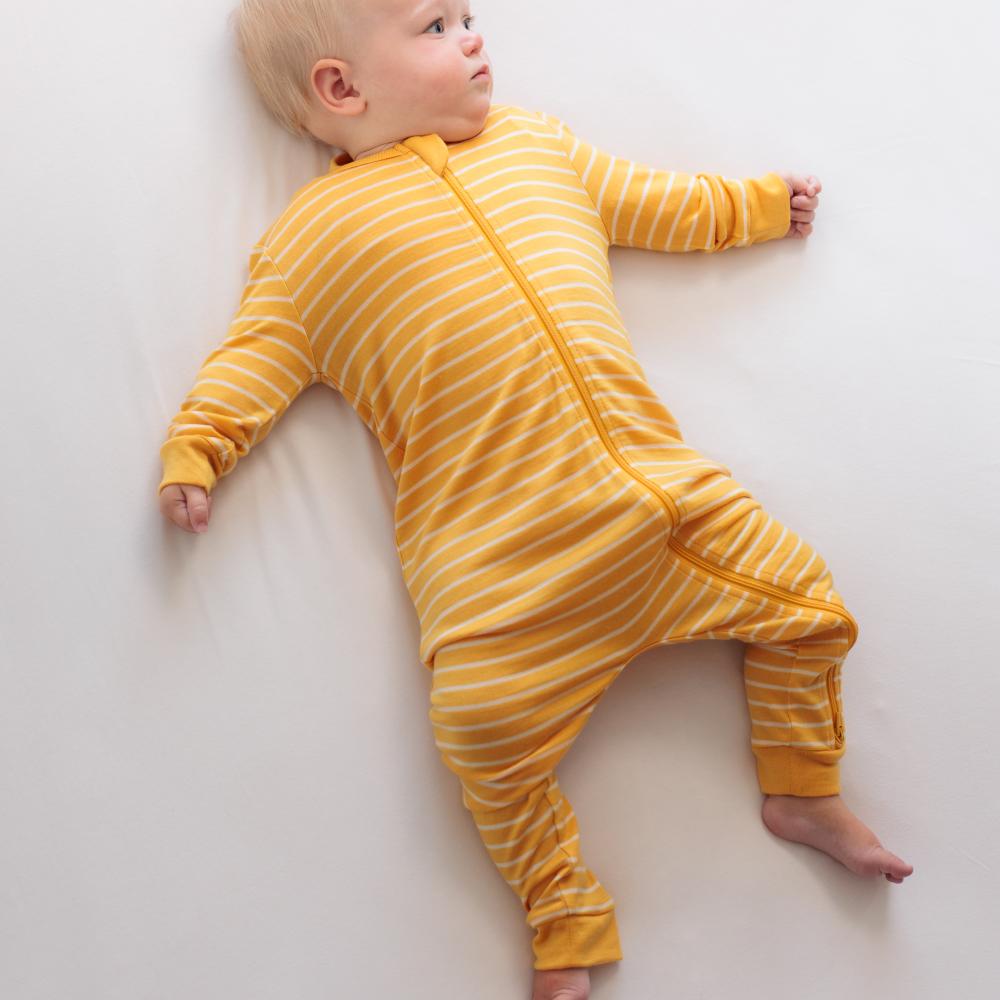 A baby lying on a light gray surface wearing a yellow and white striped jersey merino footless sleeper with a front zipper.