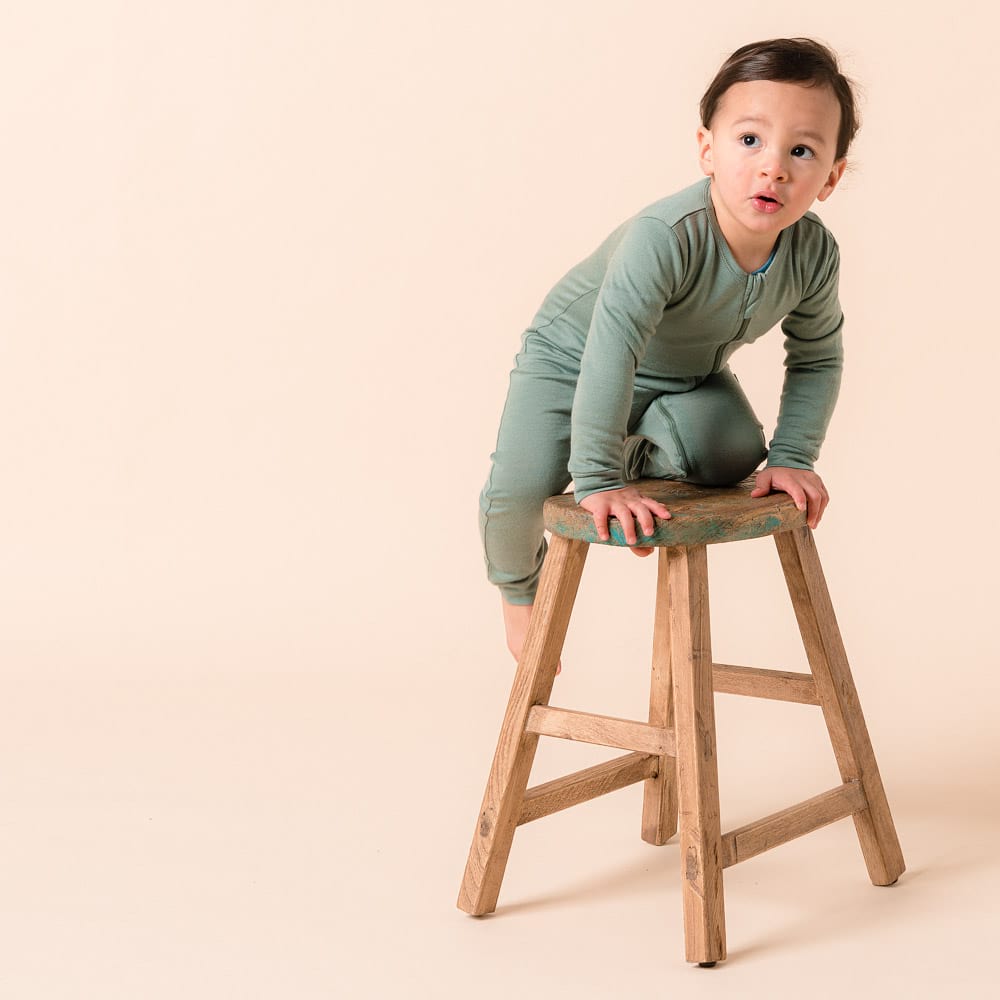 Child in a merino onesie sitting on a wooden stool against a beige background