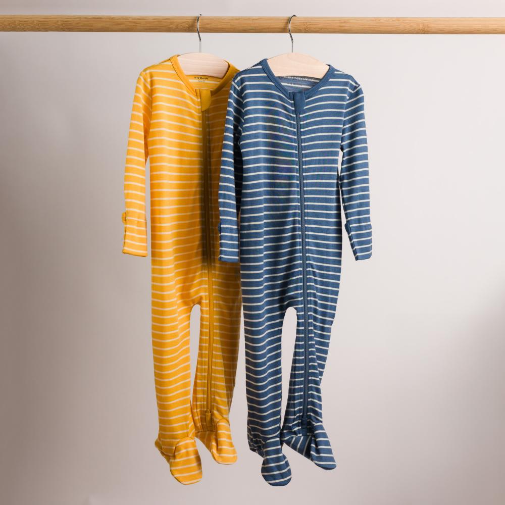 Two long-sleeve footed zip-up baby pajamas hang on wooden hangers from a wooden rod against a plain light background. The pajama on the left is mustard yellow with thin white horizontal stripes, and the pajama on the right is slate blue with thin white horizontal stripes.