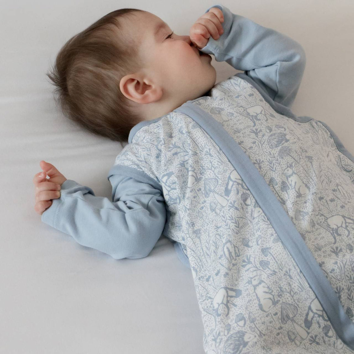 A baby lying down wearing a blue baby sleeping bag with woodlands print with hand up to their mouth.