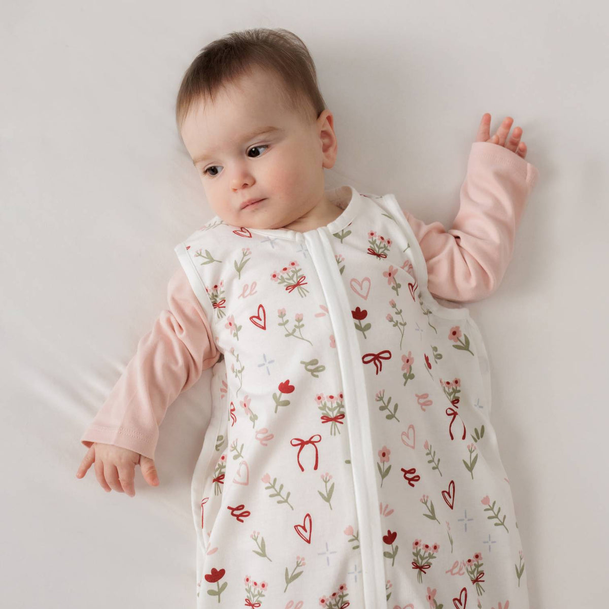 A baby lying down wearing a cotton sleeping bag with an all over bows, flowers and hearts print . 