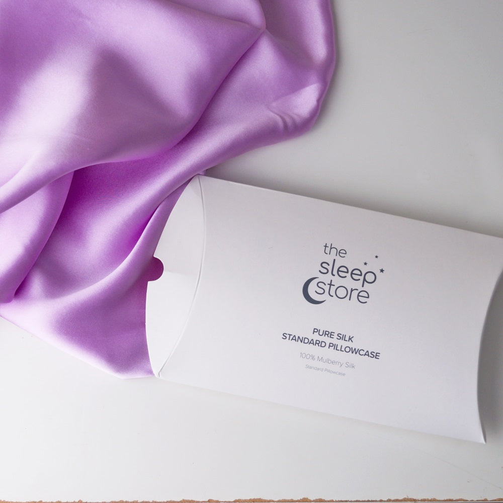 A white pillowcase packaging box printed with 'the sleep store' and 'PURE SILK STANDARD PILLOWCASE' lies on a white surface next to draped lavender-purple satin fabric.