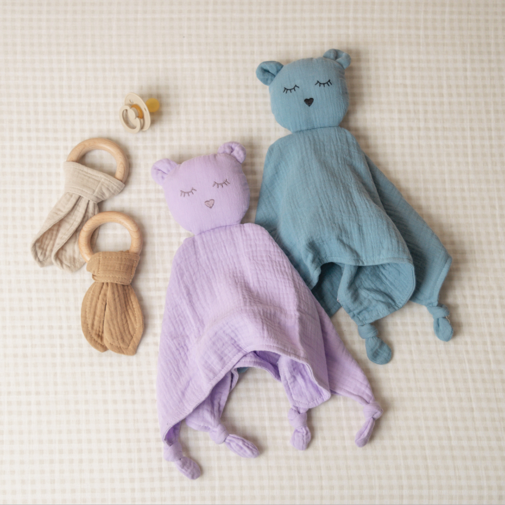 Two soft baby toys, one purple and one blue, on a textured surface with two wooden rings.