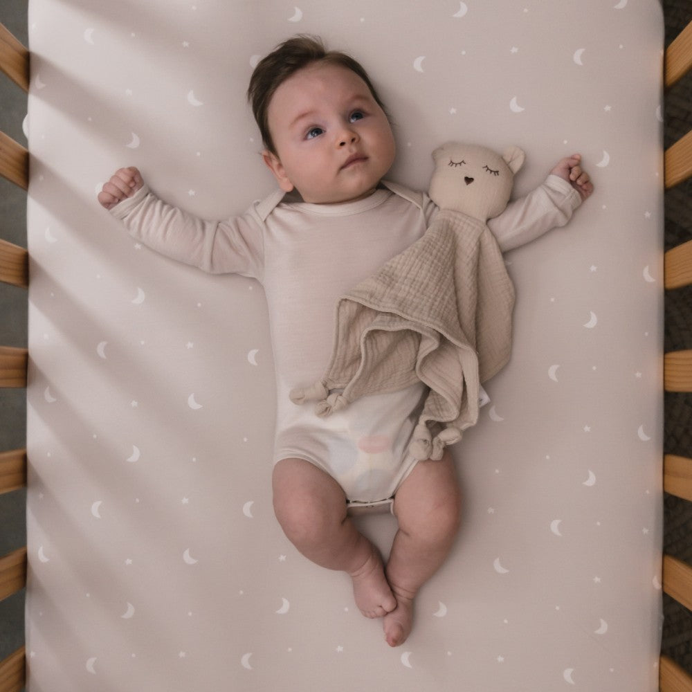 Baby lying on a crib with a teddy bear cotton muslin comforter surrounded on a patterned sheet.