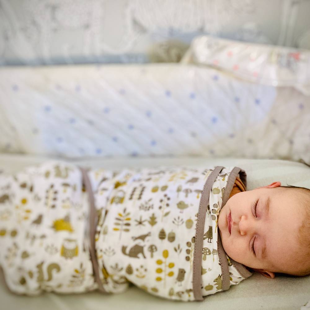Newborn baby sleeping in a white bassinet with a white and brown/green forest patterned miracle swaddle blanket