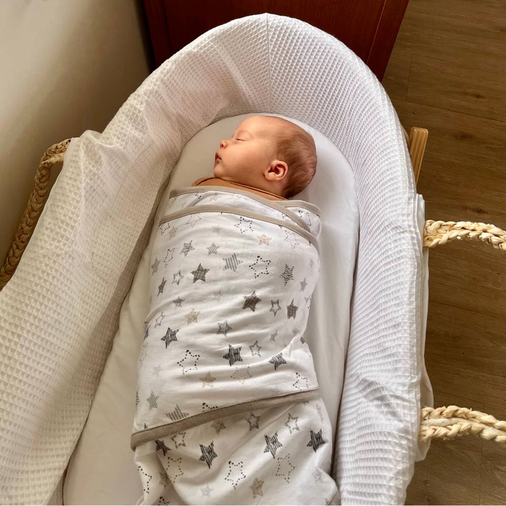 Newborn baby swaddled in a white/grey star miracle blanket lying in a white crib.