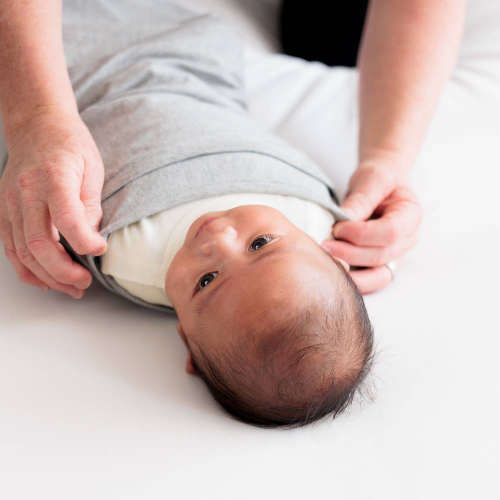 Baby being swaddled in a grey miracle blanket by an adult on a white surface