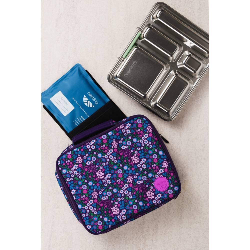 A floral patterned lunch bag in purple, blue, and pink lies on a light beige surface, alongside a blue ice pack and a stainless steel bento box.