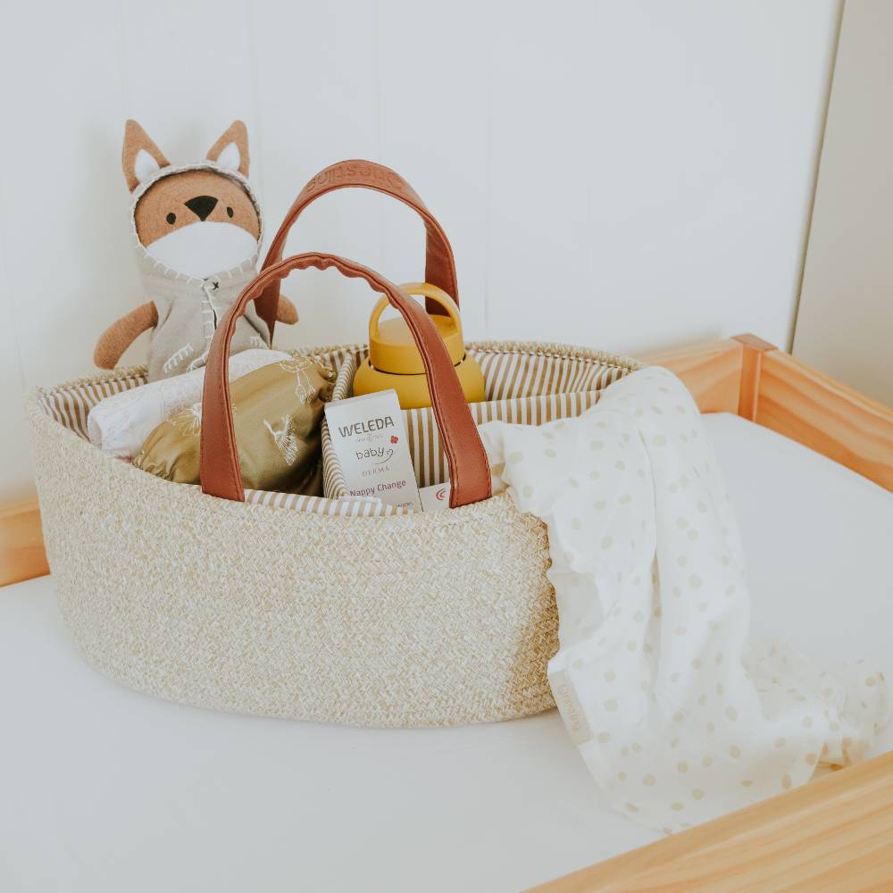 Beige Baby basket with tan faux leather handles with toys and a blanket on a white surface