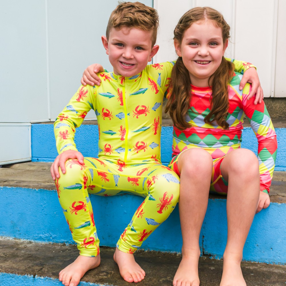 Two children sitting on a blue step wearing colorful swimsuits.