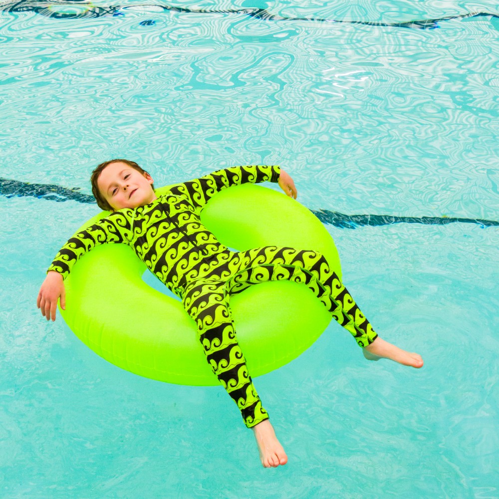 Child in a green wavy-patterned swimsuit lying on a green inner tube in a pool.