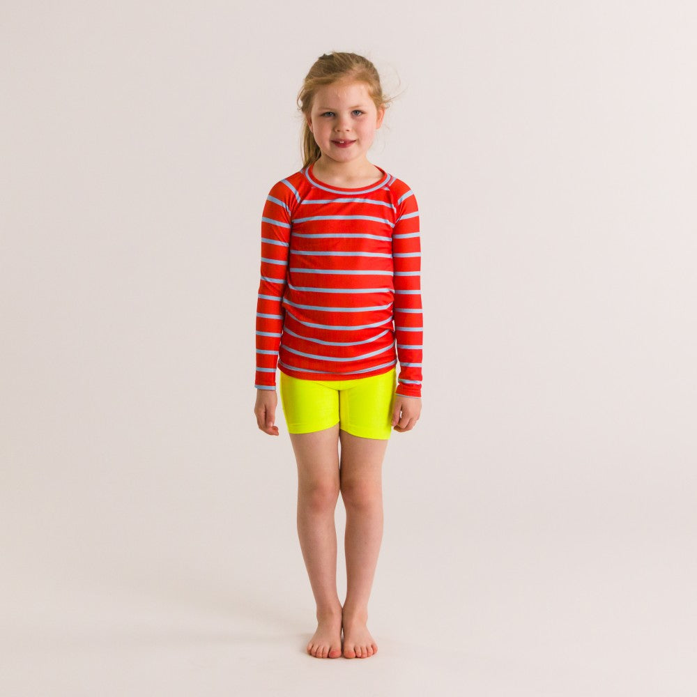 Young girl wearing a red striped rash top and yellow shorts on a plain background
