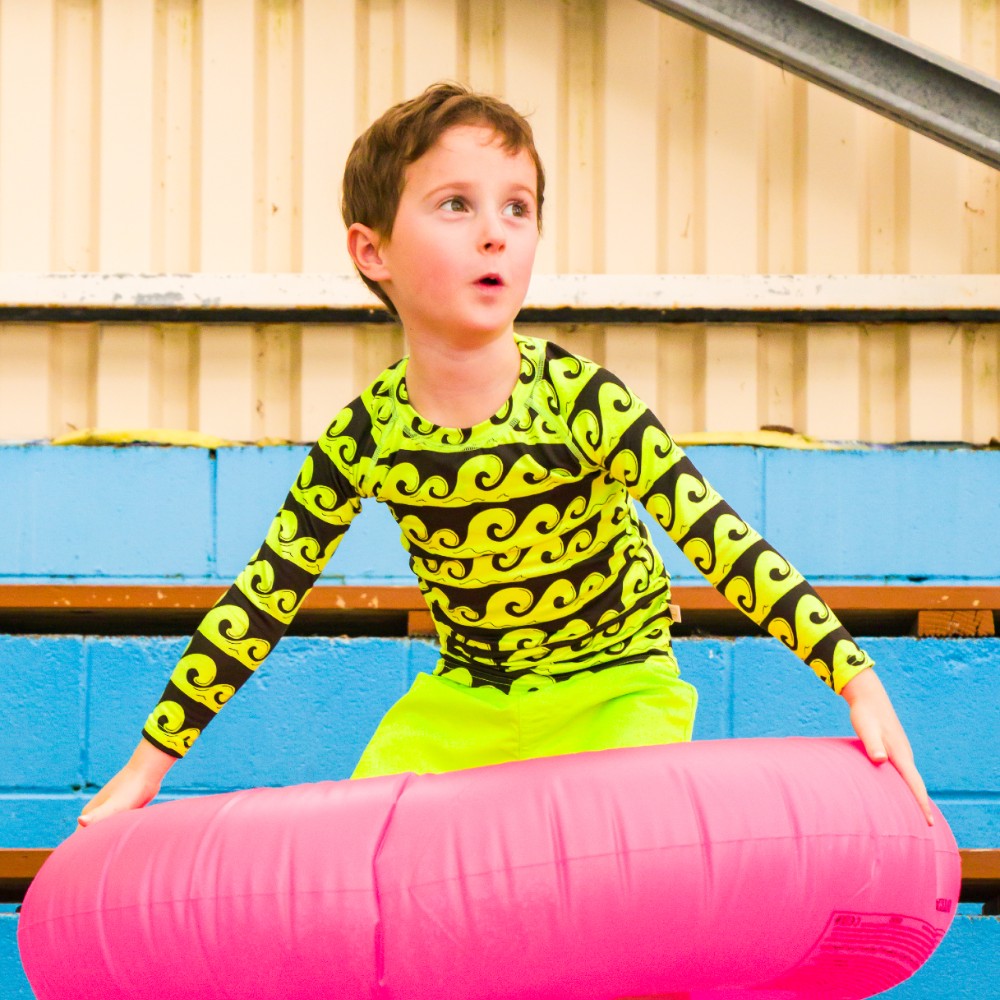 Child wearing a green and black wave rash top and green shorts sitting on a pink inner tube with a blue wall and wooden bench in the background.