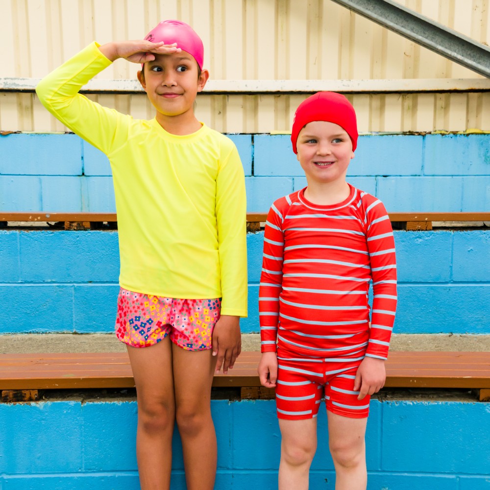 Two children in colorful swim gear standing by a pool.