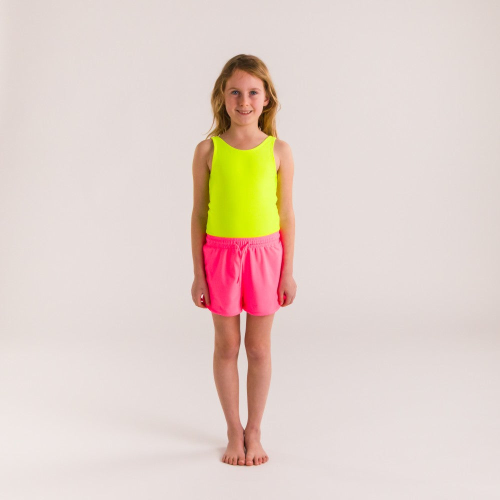 Young girl wearing a bright neon yellow swimsuit and neon pink board shorts on a plain background