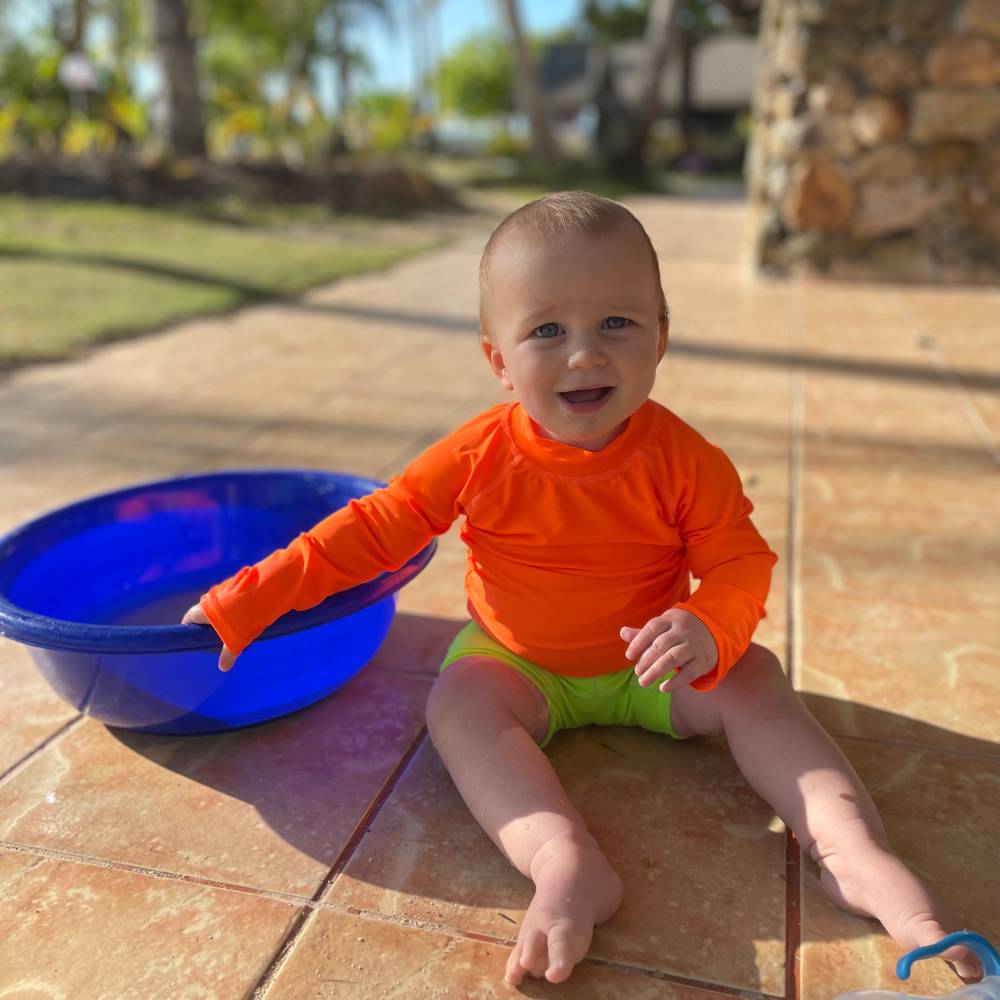 Child in orange rash vest and neon green swim shorts sitting on a tiled floor next to a blue plastic tub outdoors.