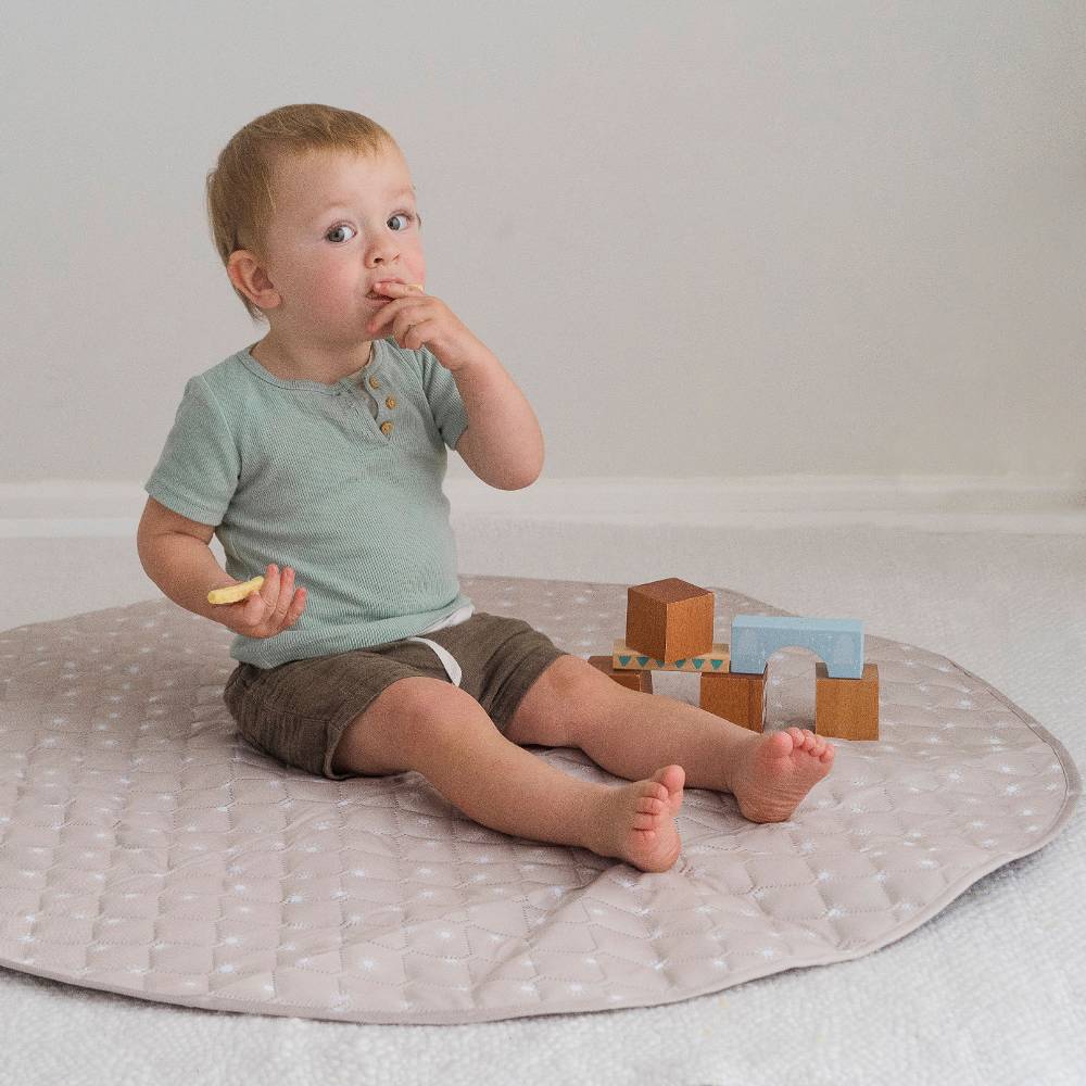 A baby sitting on a round quilted play mat, wearing a sage green t-shirt and brown shorts, with wooden blocks nearby on a light-colored floor.