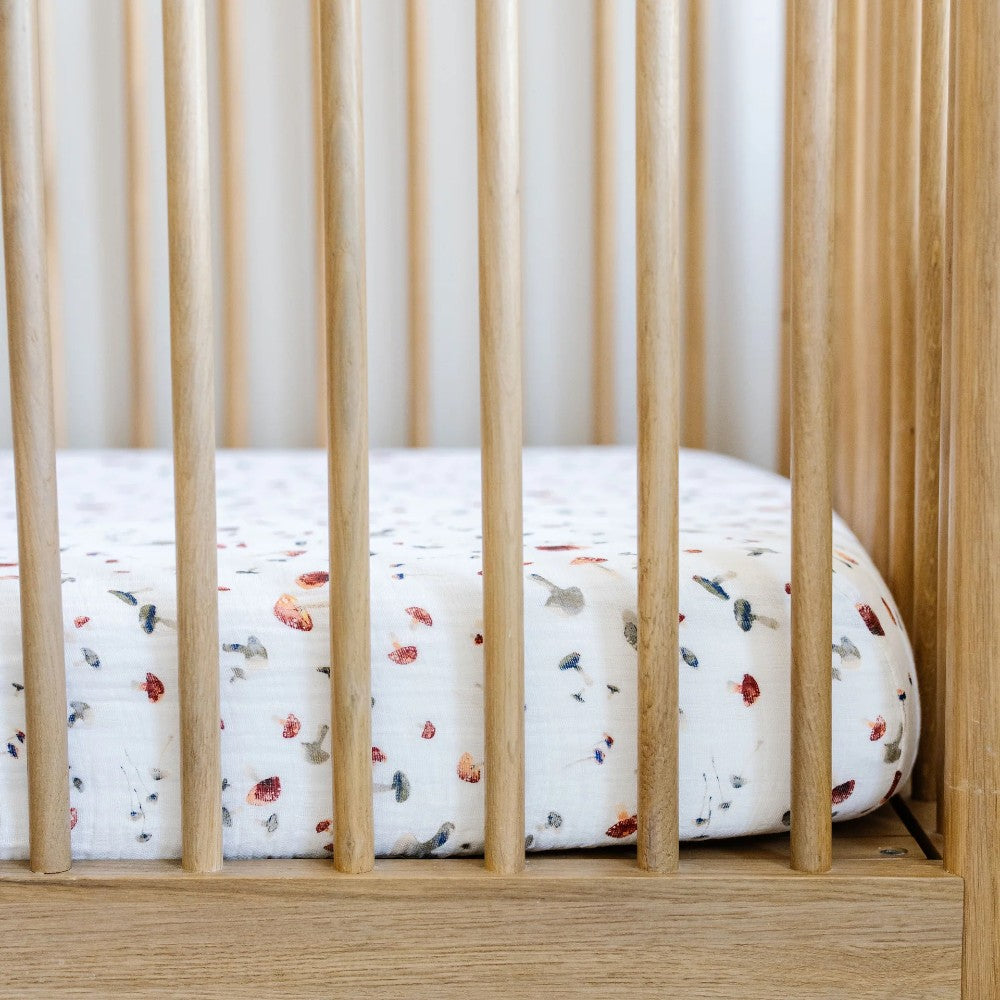 A white cotton muslin fitted sheet with mushroom pattern on a wooden cot with a blurred background