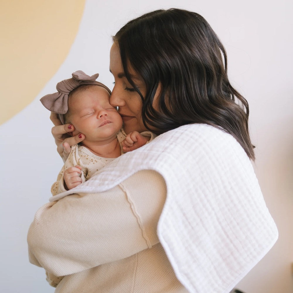 Woman holding a newborn baby with a purple bow with a white cotton muslin burp cloth over her shoulder with a soft background