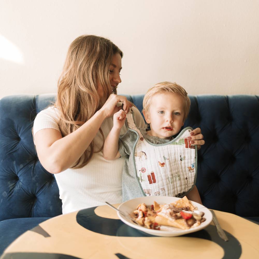 Woman and child sitting on a blue couch with a plate of food in front of them as she places the bib around the childs front