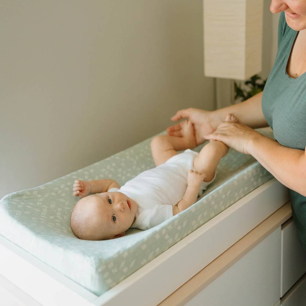 Baby lying on a green and white patterned changing pad cover on a white changing table with a woman assisting