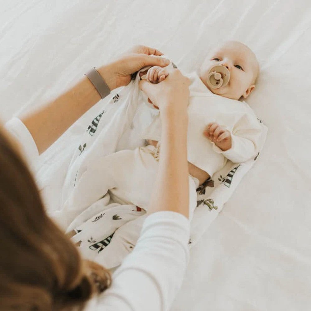 Baby being put into a forest animal themed muslin sleeping bag by an adult on a white blanket