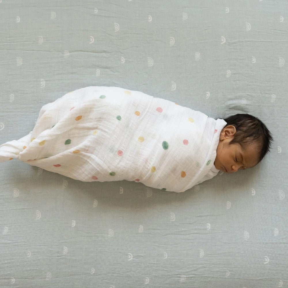 Newborn baby swaddled in a white blanket with colorful polka dots on a sage green patterned surface.