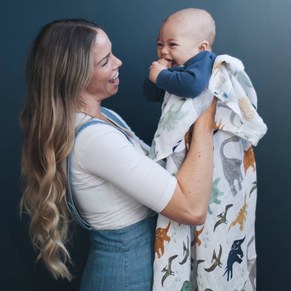 Woman holding a baby wrapped in a dinosaur-patterned muslin swaddle blanket against a dark background