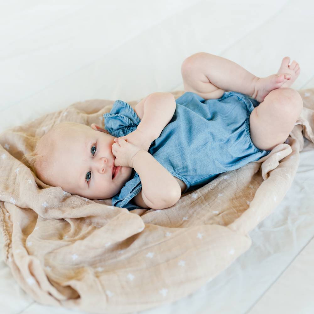 Baby lying on a beige and white cross muslin swaddle  blanket wearing a blue outfit.