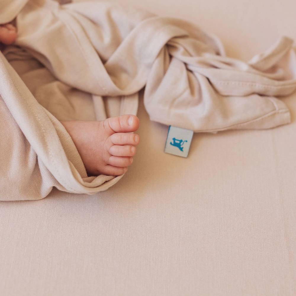 Baby's foot peeking out from under a soft dusky pink blanket on a matching sheet with a brand tag on a light background