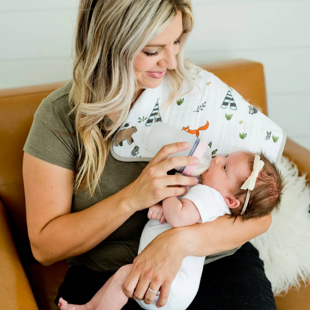 Woman holding a baby and feeding them with a bottle on a couch with a white forest animal patterned burp cloth over her shoulder 