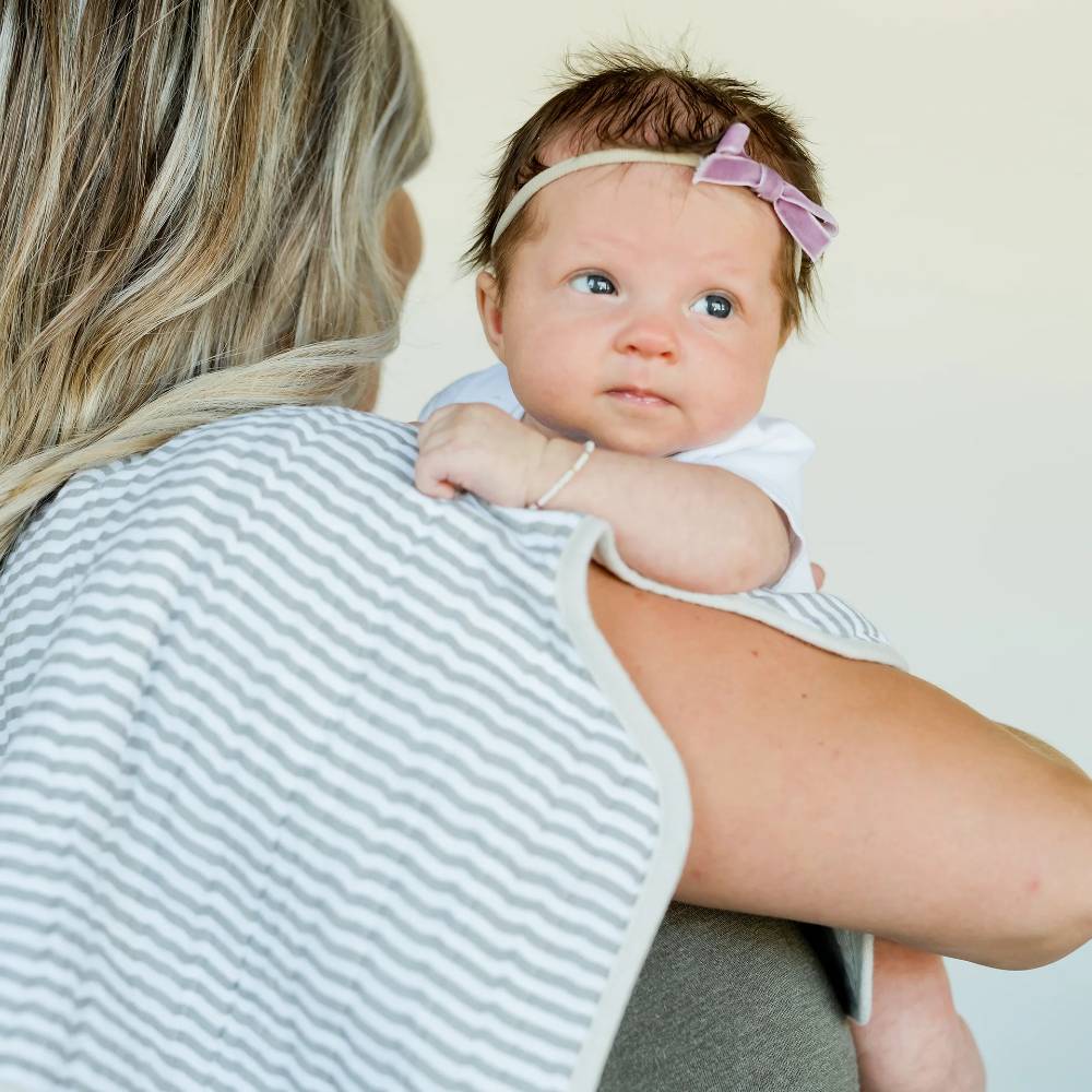 Woman holding a baby with a grey and white burp cloth on shoulder against a plain background