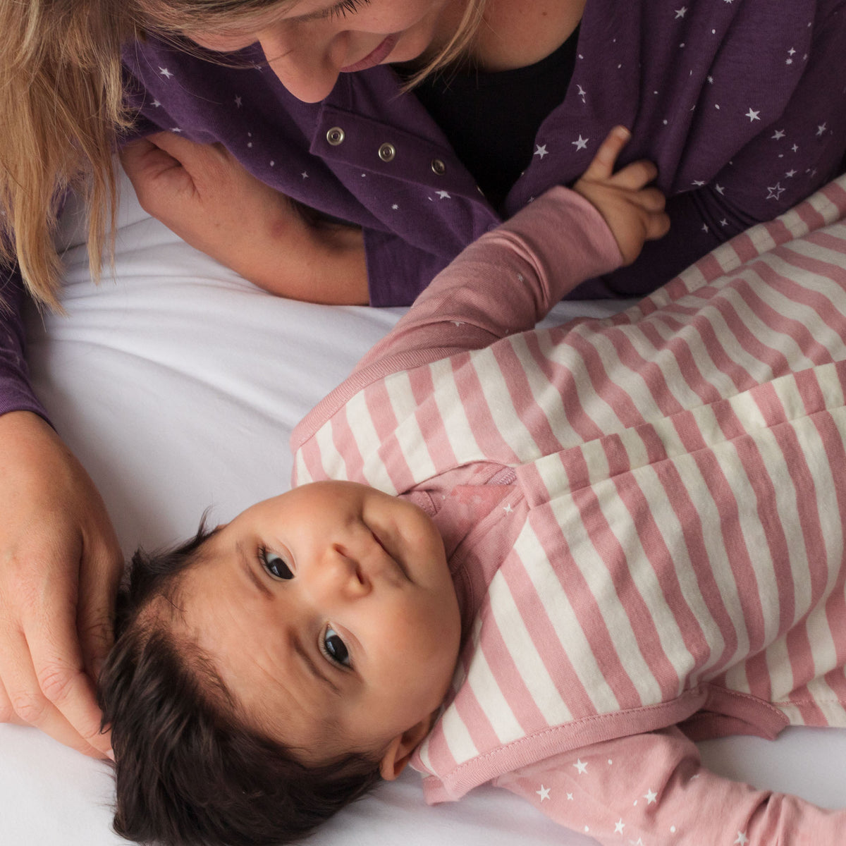 Baby in pink and white striped outfit lying on a white surface with a person in a purple jacket.