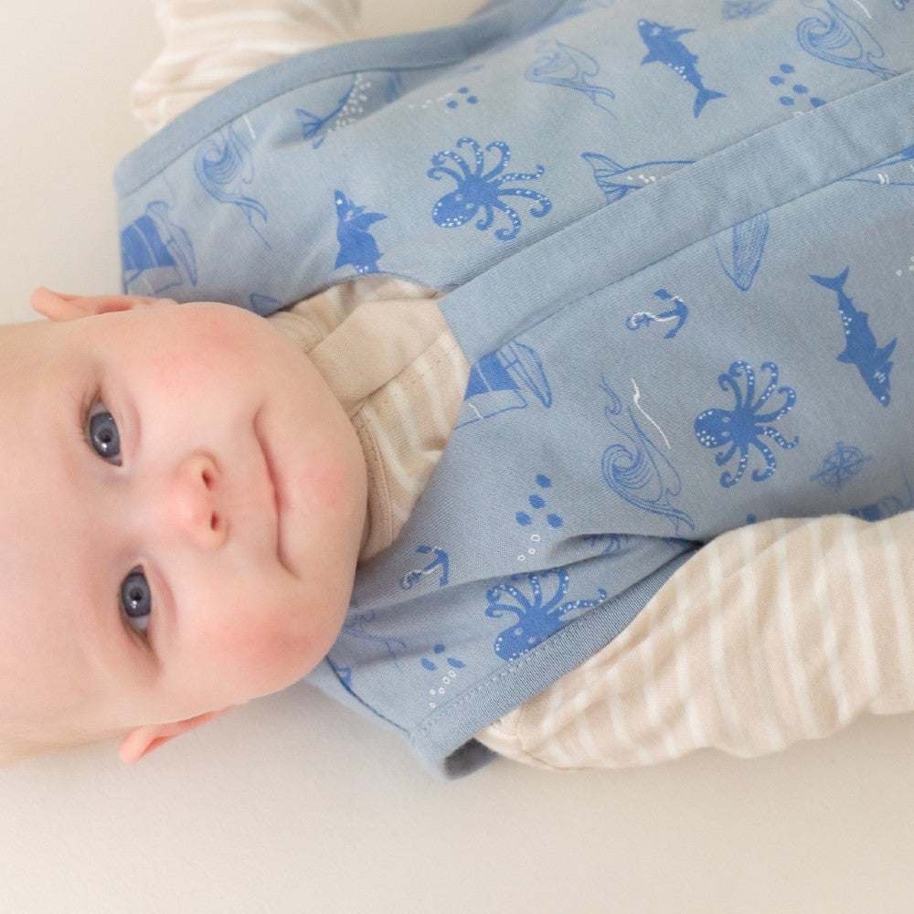 Baby lying on a white surface with a blue bag wrap featuring sea creature patterns.