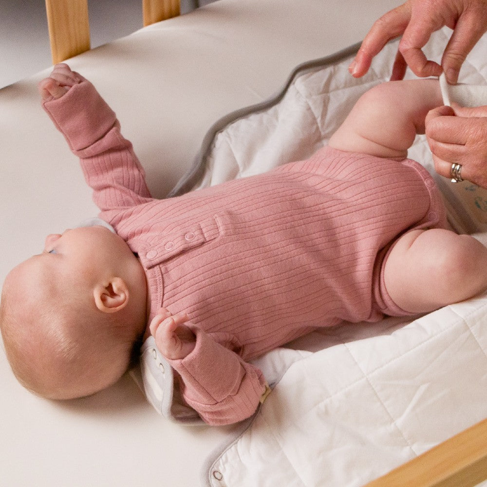 Baby in a pink outfit lying on a white blanket with a person's hand visible.