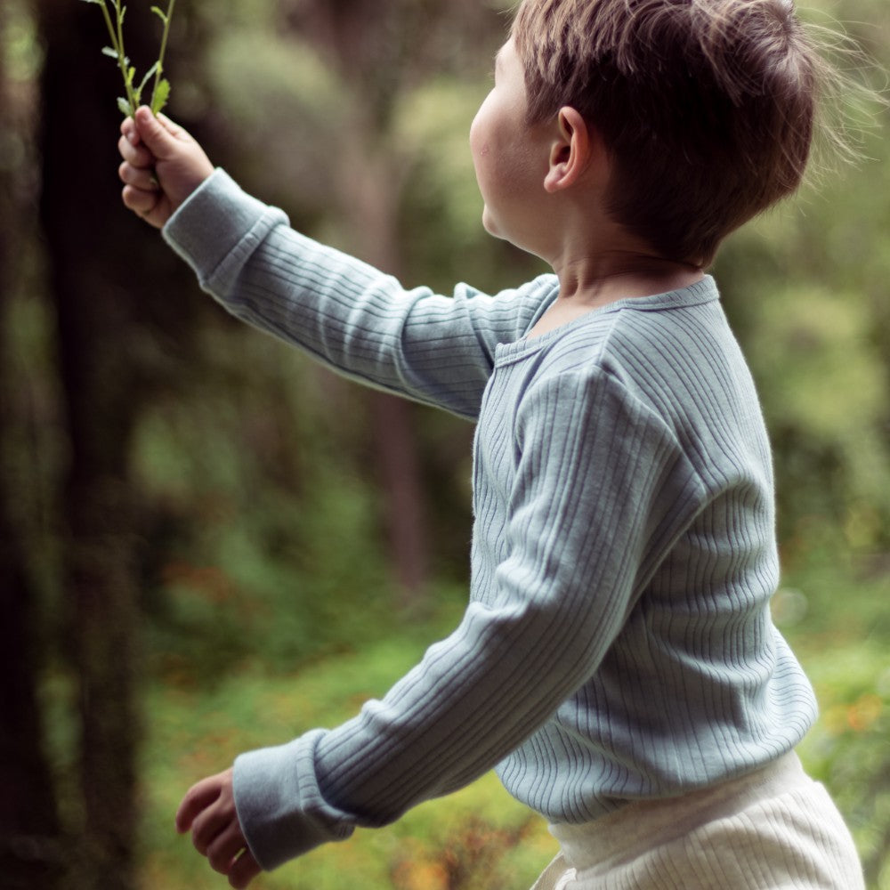 Child in a light blue rib bodysuit holding a leaf in a forest setting