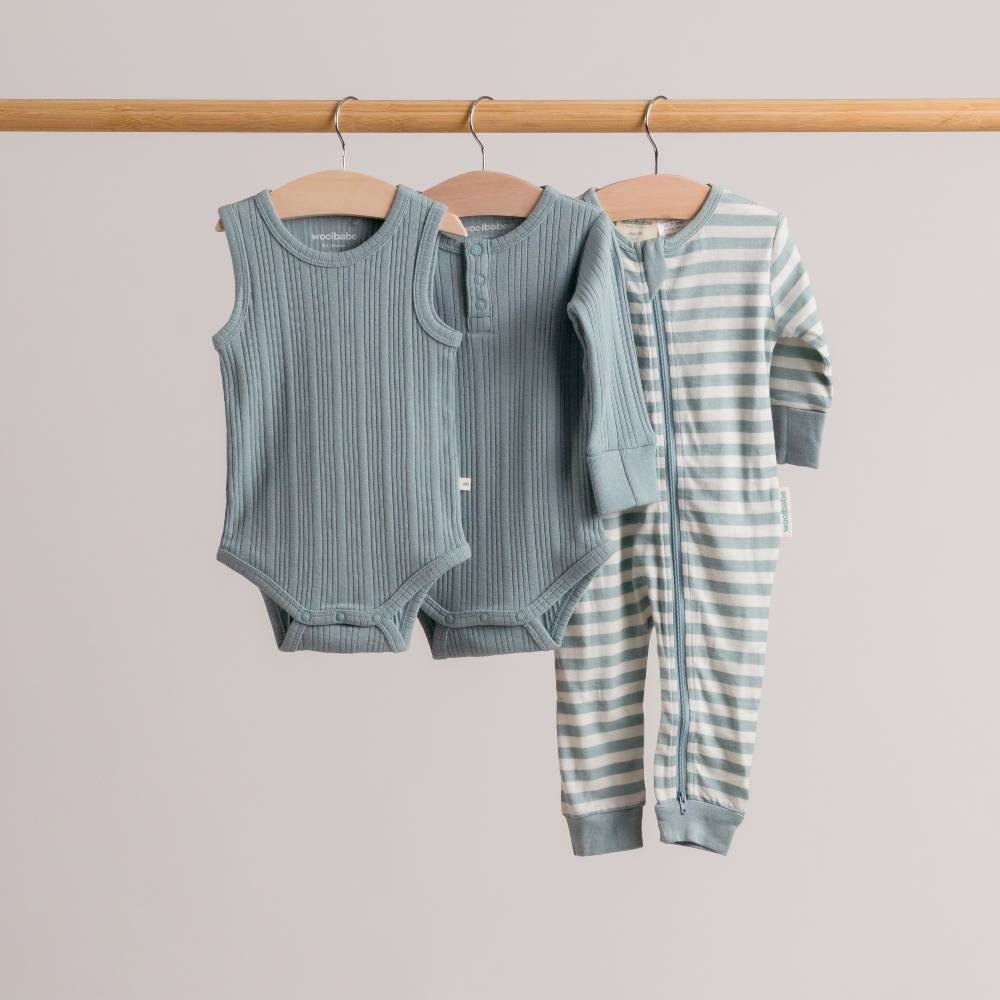 Children's clothing set with a woolbabe rib bodysuit, long-sleeve shirt, and striped pants on hangers against a neutral background.
