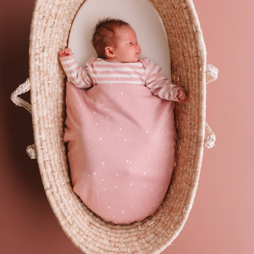 Newborn baby in a pink outfit and blanket lying in a woven basket on a pink background