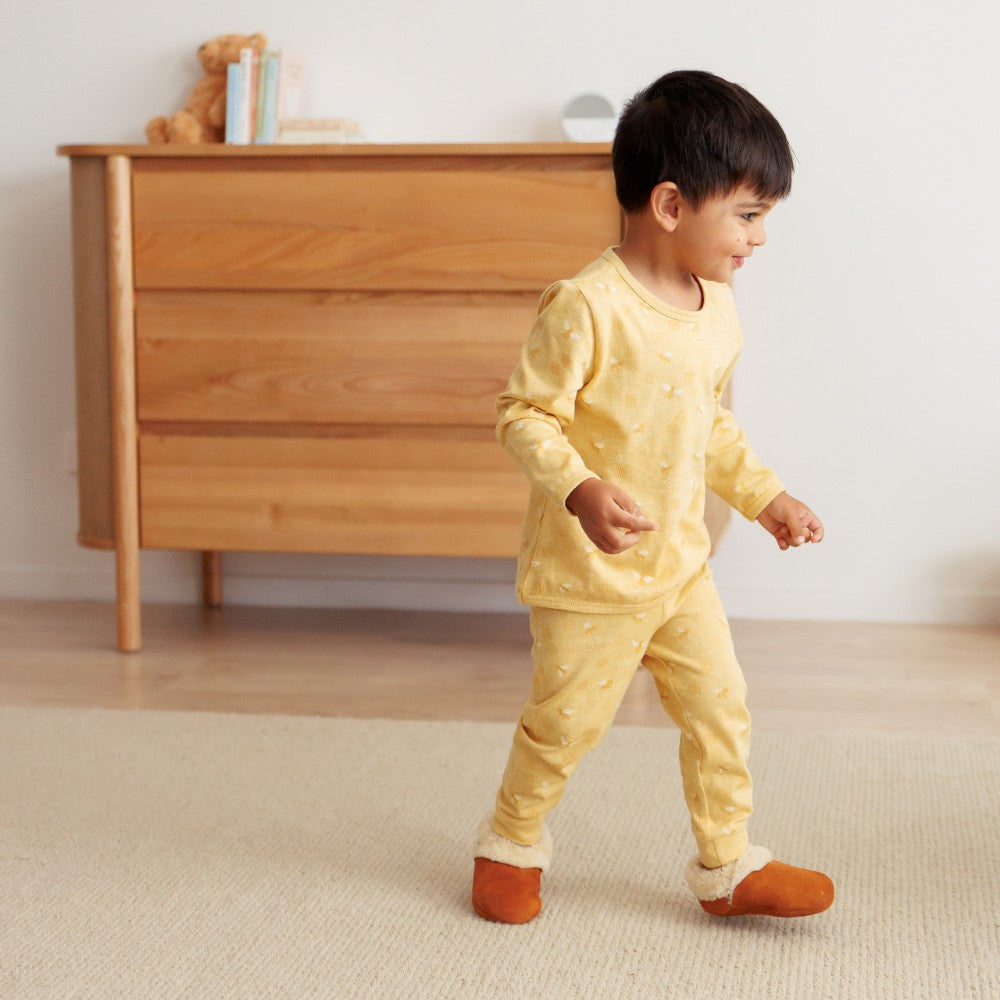 Child in yellow pajamas standing in a room with wooden furniture.