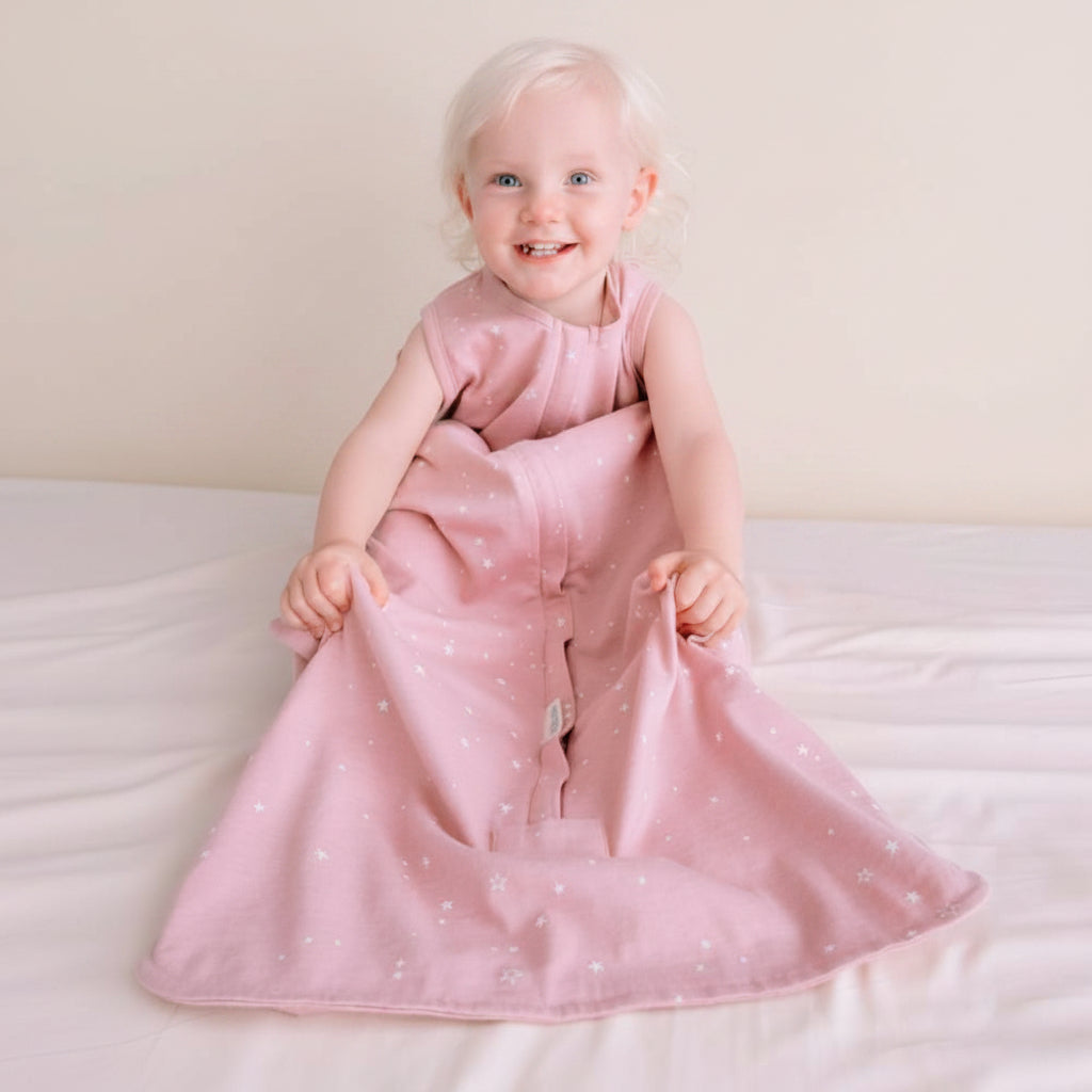 Child wearing a pink sleeping bag with white polka dots sitting on a white surface against a blue wall.