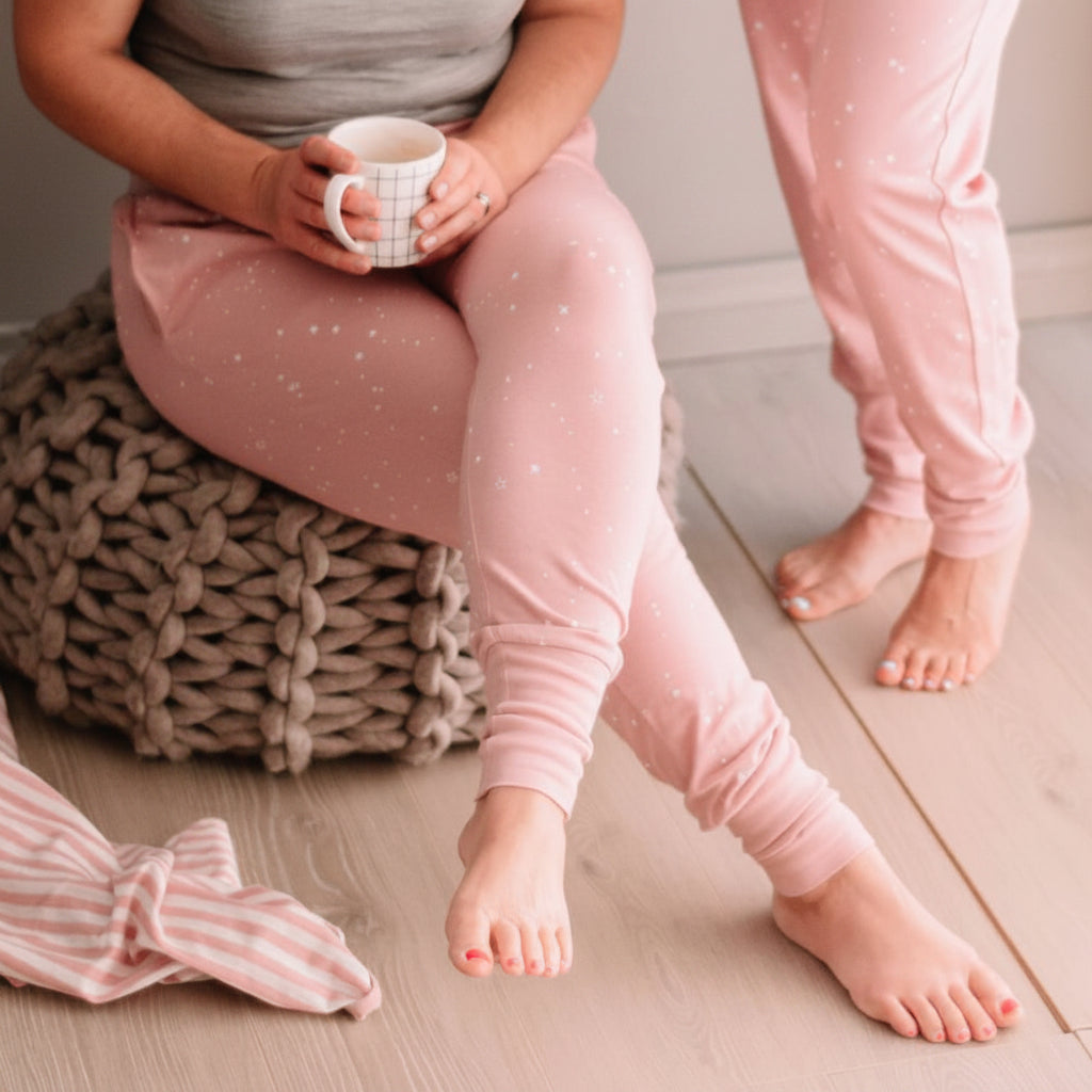 Person wearing pink pajama pants sitting on a wooden floor holding a mug.