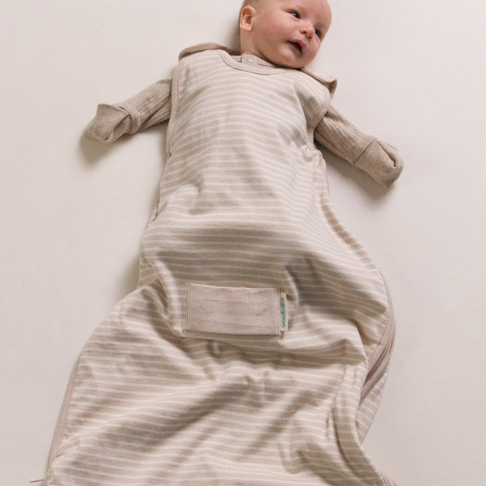 Baby lying on a plain surface wearing a beige striped baby grow with a visible brand label.