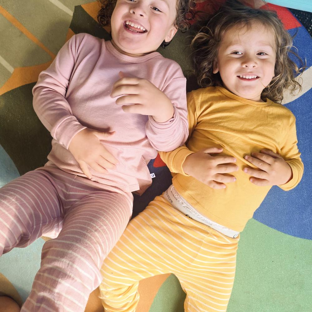 Two young children lying on a colorful floor mat, wearing matching long-sleeve jersey merino pajamas. The child on the left wears a light pink pajama set with thin white striped pants, and the child on the right wears a mustard yellow pajama set with thin white striped pants