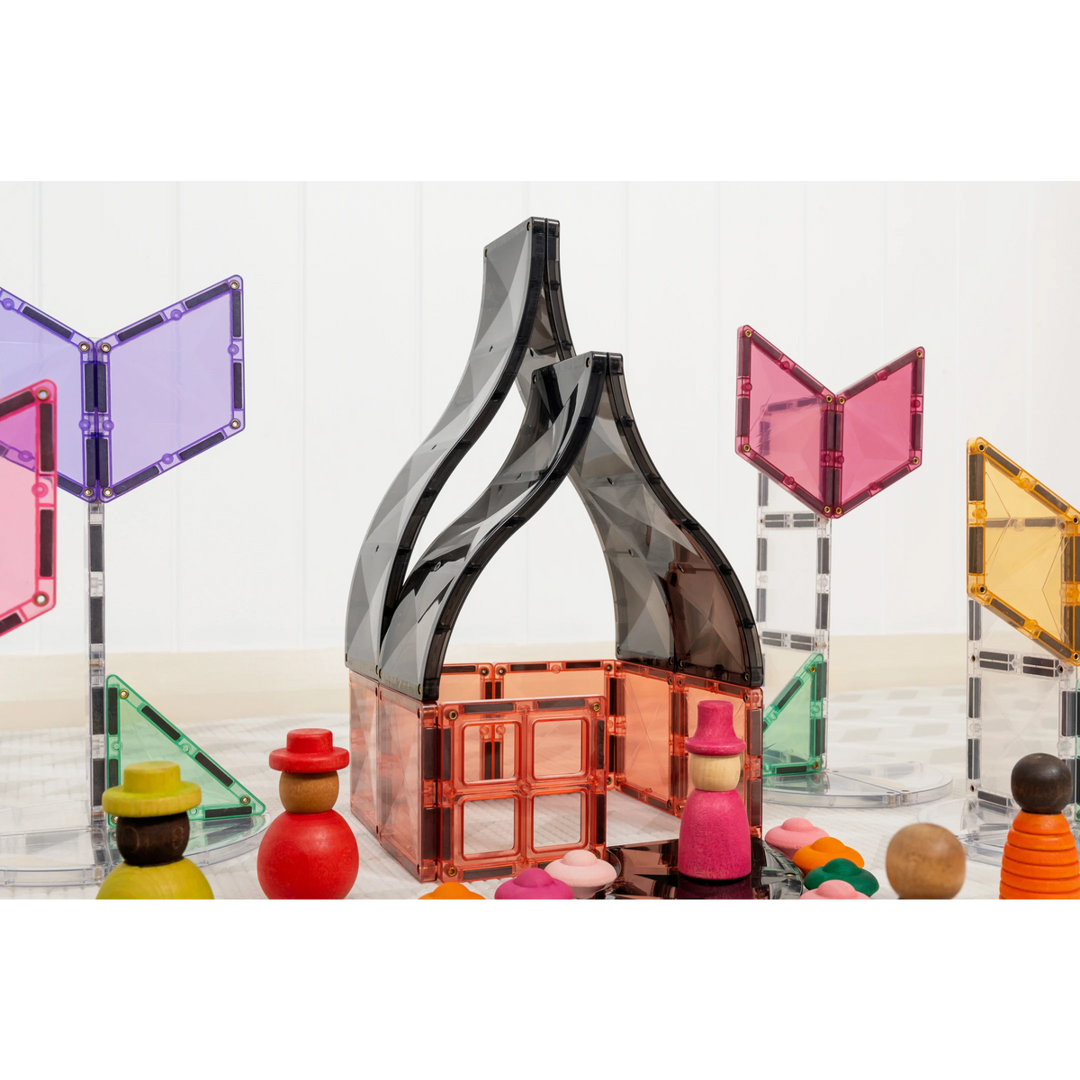Magnetic building blocks set with a translucent house structure in the center, surrounded by colorful wooden toy figures and geometric tiles on a white background.