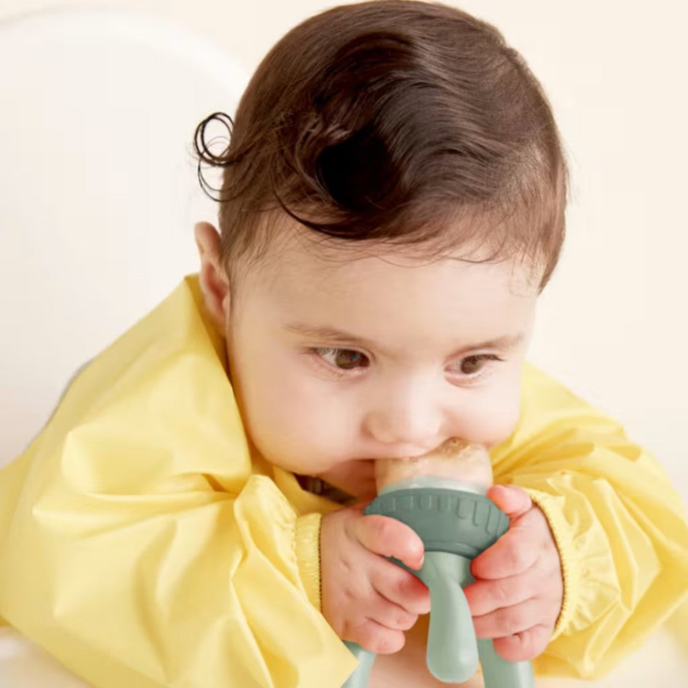 Baby in a yellow outfit holding a green feeder against a white background