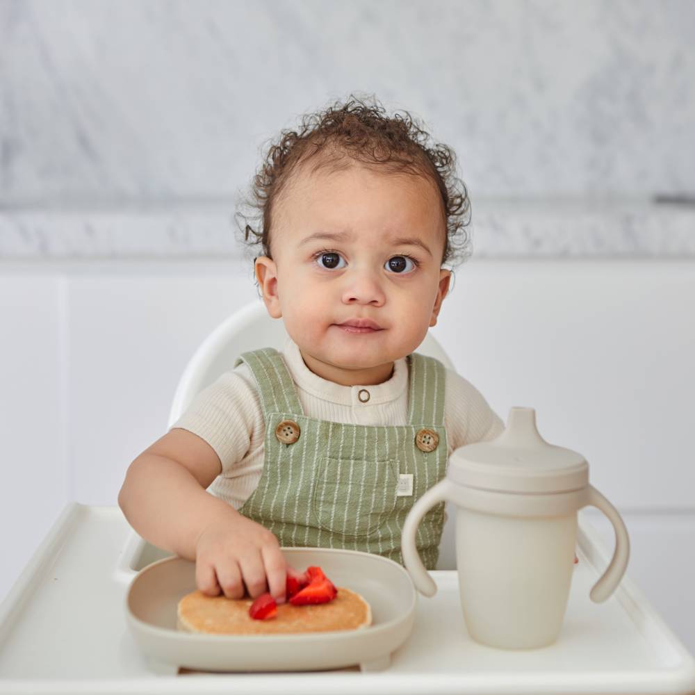 Baby in a high chair with a plate of food and a sippy cup on a white background