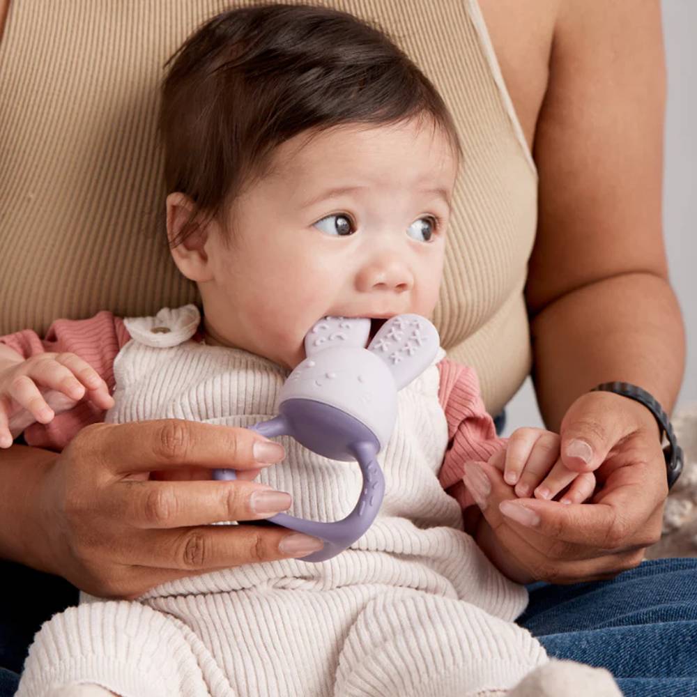 Baby holding a purple teething toy with a person holding the baby's hands