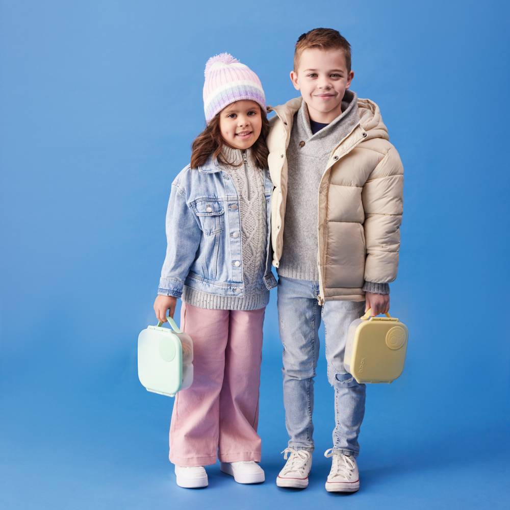 Two children standing together holding mini lunchboxes against a blue background