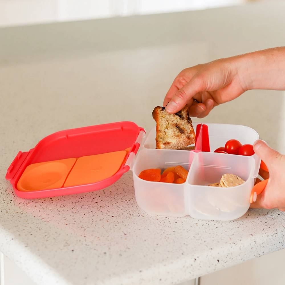 Person placing a sandwich into a b.box mini lunchbox with compartments for different food items on a countertop.