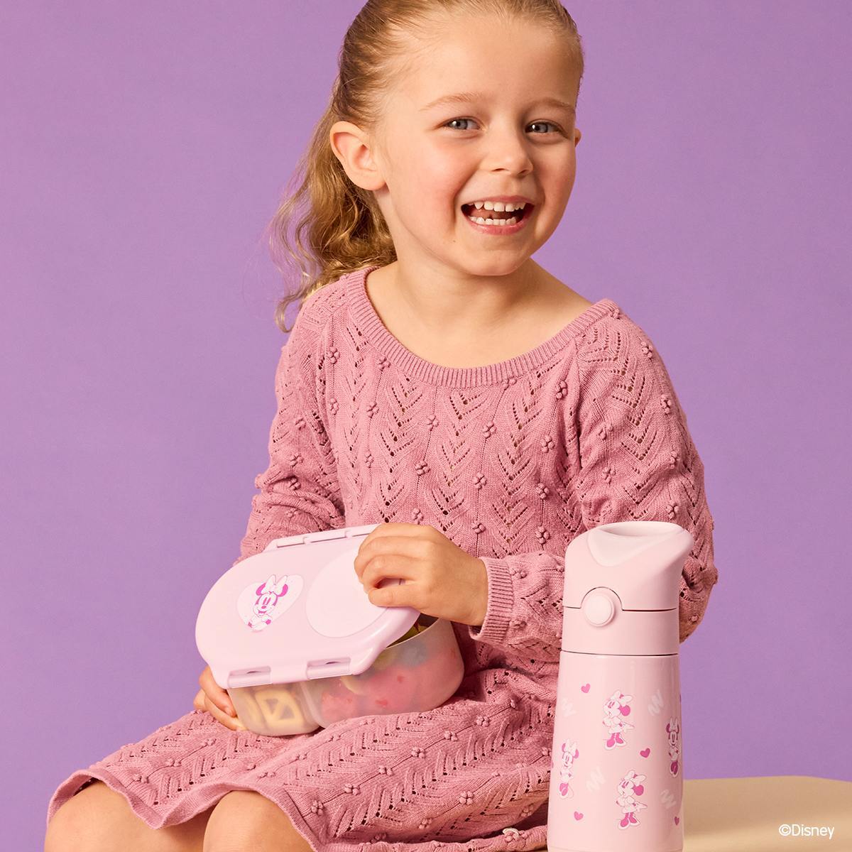 A young child smiling, sitting down holding a pink snackbox with a matching drink bottle next to them. 